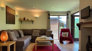 A living room with a brown sofa, a wooden coffee table, a red rug, a red armchair, and a television on the wall. - Water Mill Vacations, Badgers Oak (Calbourne)