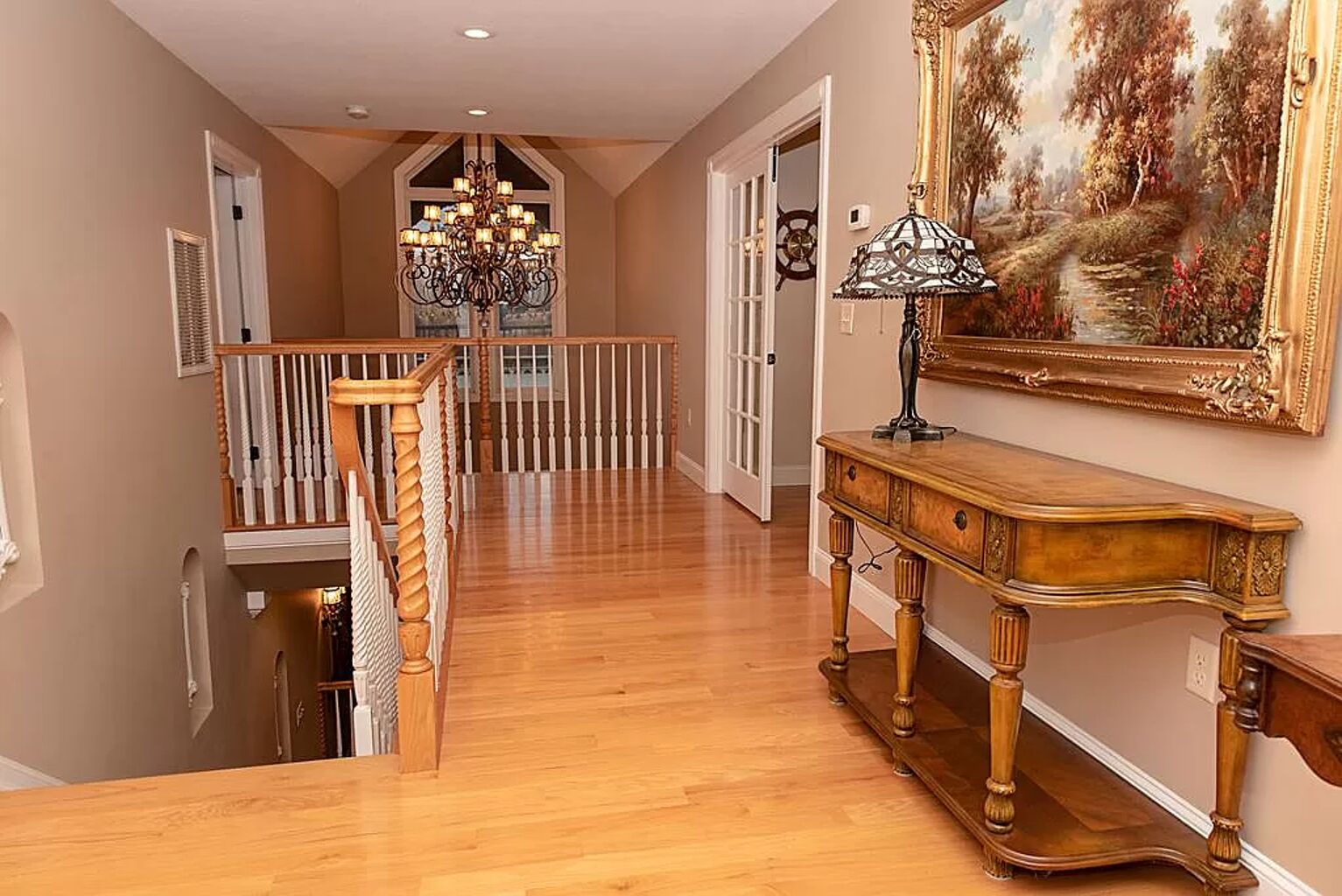 A spacious hallway with wooden flooring, a staircase with a railing, a chandelier, and a painting on the wall.