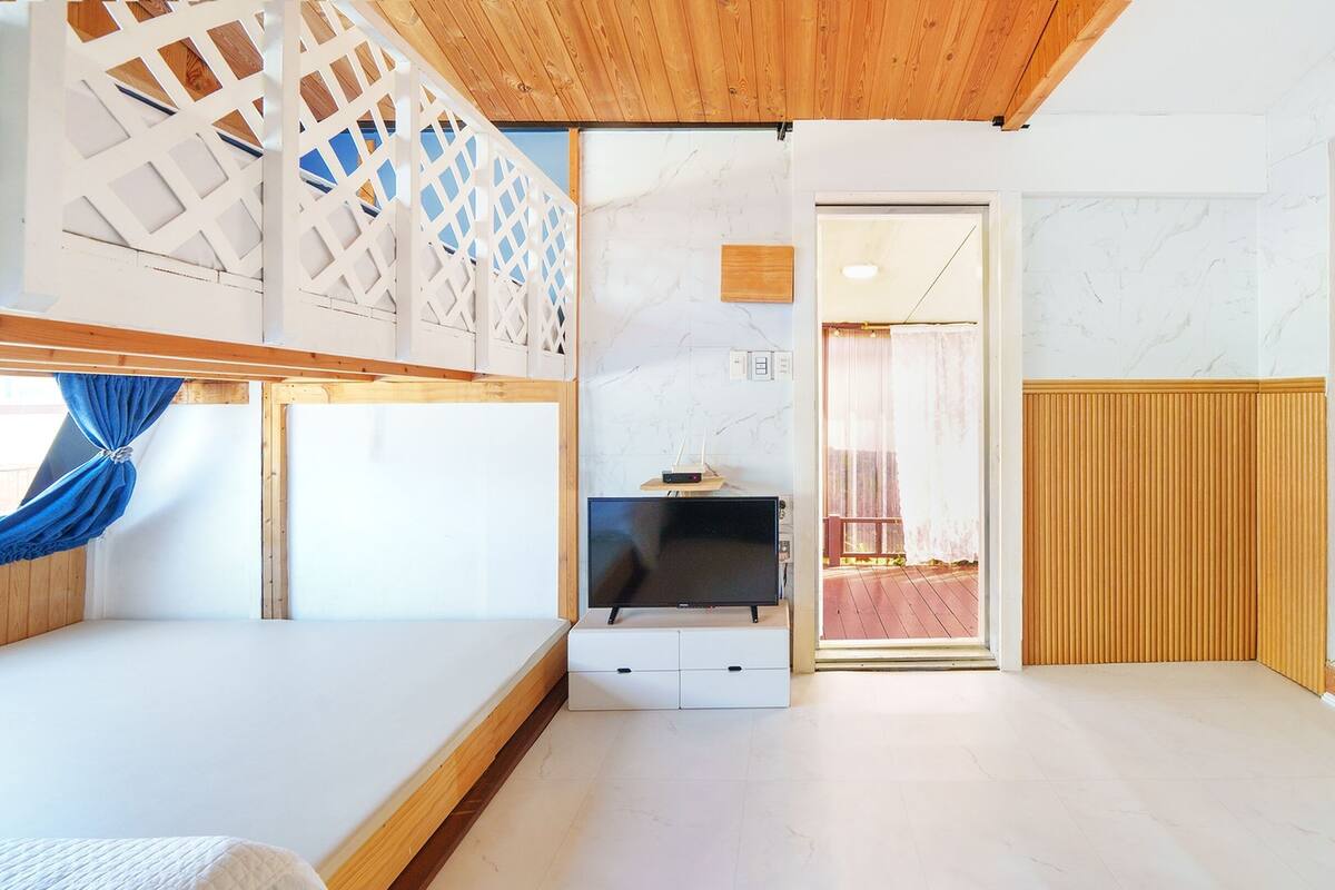 A bedroom with a bed, a TV, a wooden ceiling, a white wall, and a door leading to another room.