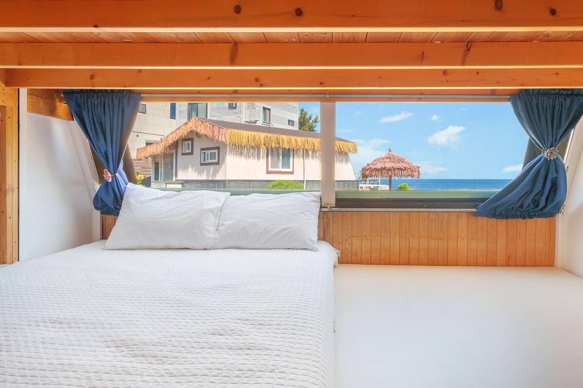 A bedroom with a bed, wooden ceiling, and a view of a beach and buildings.