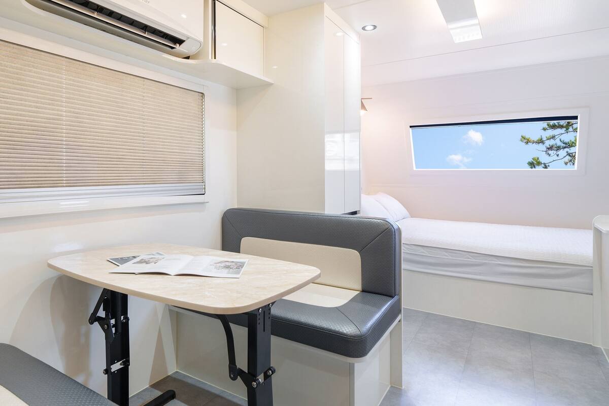 A compact, modern room with a bed, a small table with a magazine, a window with blinds, and a ceiling fan.