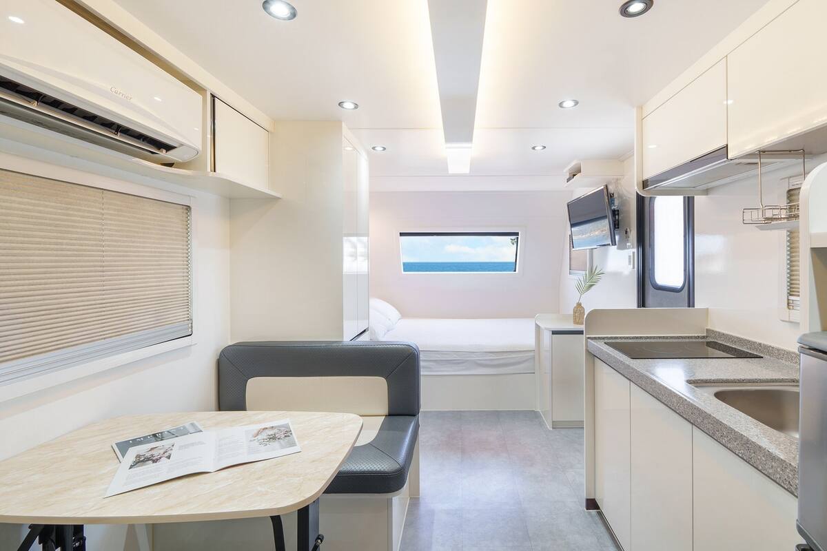 A compact kitchenette with a small dining area, a bed with a window, and a TV mounted above the bed.