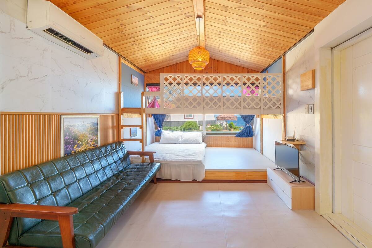 A room with a wooden ceiling, a bunk bed, a green leather sofa, a TV, and a view of the sea.