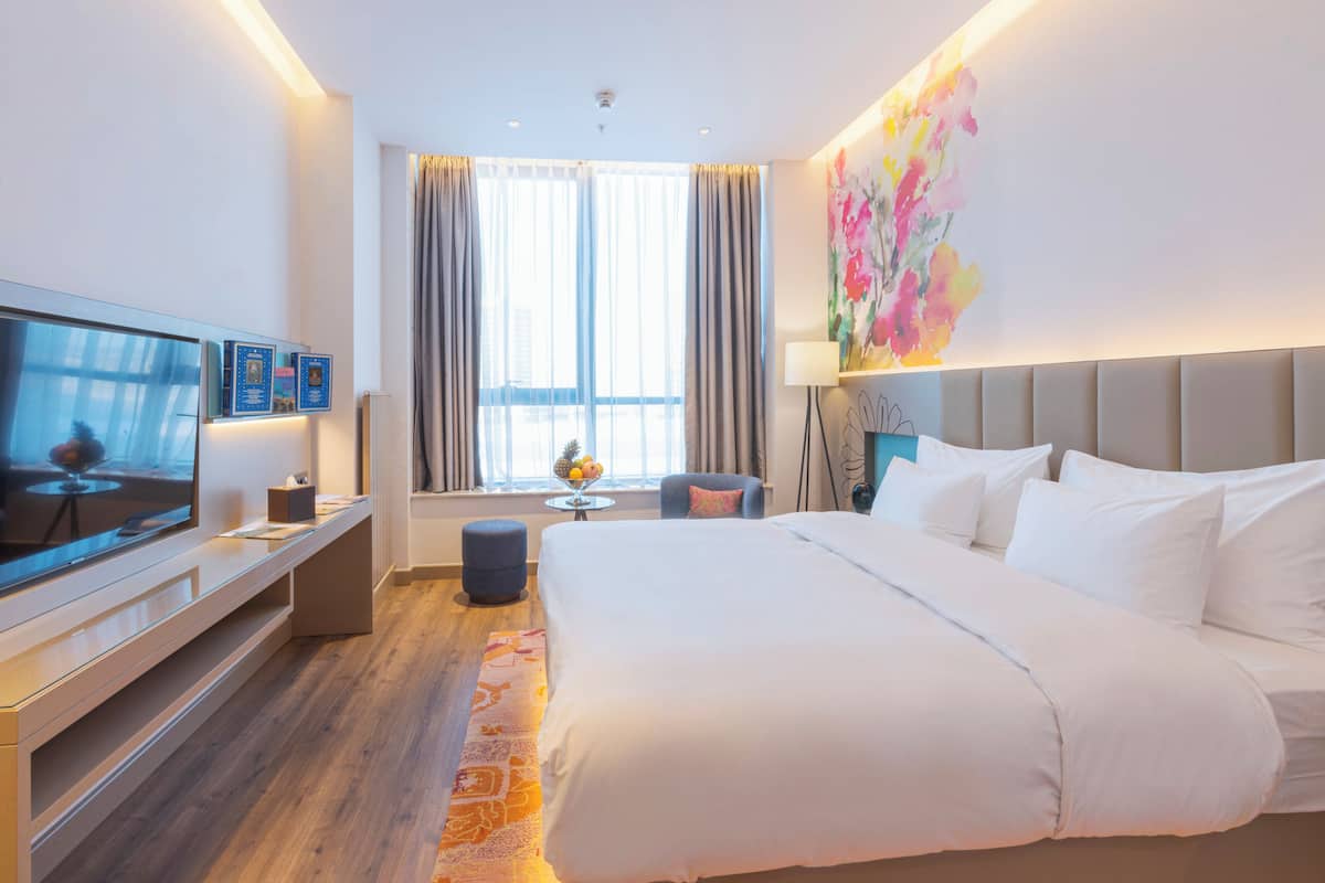 A modern hotel room with a large bed, a flat-screen TV, a desk with a chair, a small table with a lamp, and a large window with curtains.