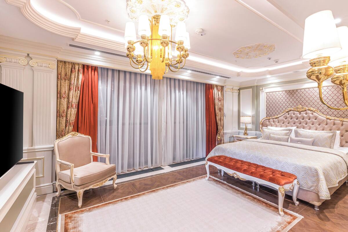 A luxurious bedroom with a large bed, a chaise lounge, a bench, a chandelier, and a television.