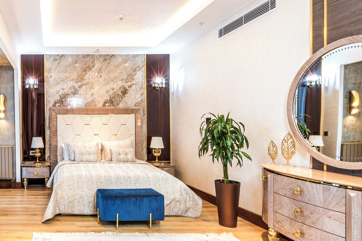 Ein Schlafzimmer mit einem großen Bett, einer blauen Bank, einem Topf mit Pflanze, einem Schminktisch und einem Spiegel.