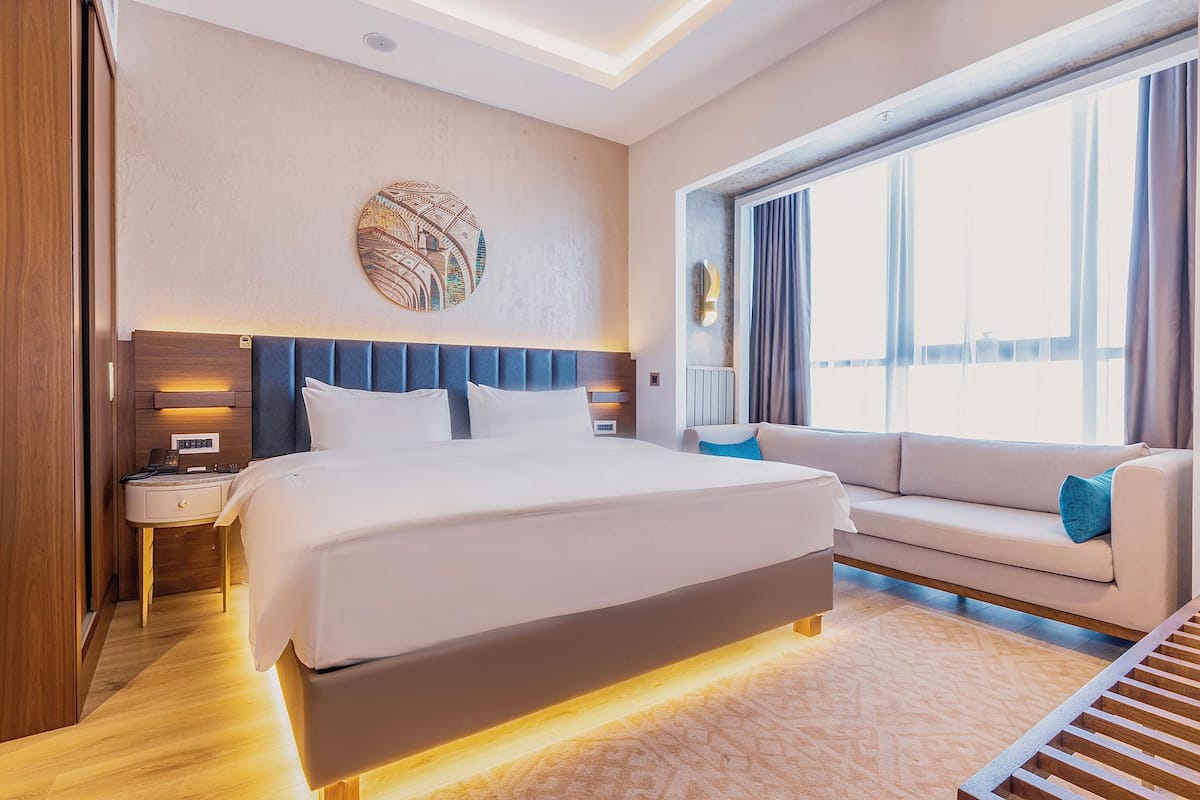 A modern hotel room with a large bed, a sofa, a round wall decoration, and a large window with curtains.