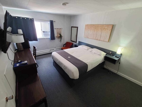 A hotel room with a bed, bedside table, lamp, desk, chair, and a window with curtains.