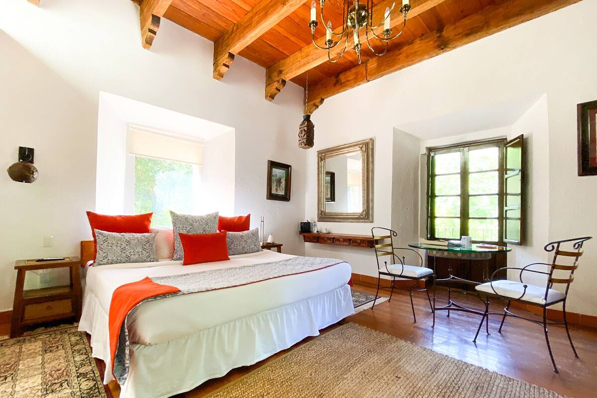 A bedroom with a large bed, a desk, and two chairs. There are windows and a chandelier.
