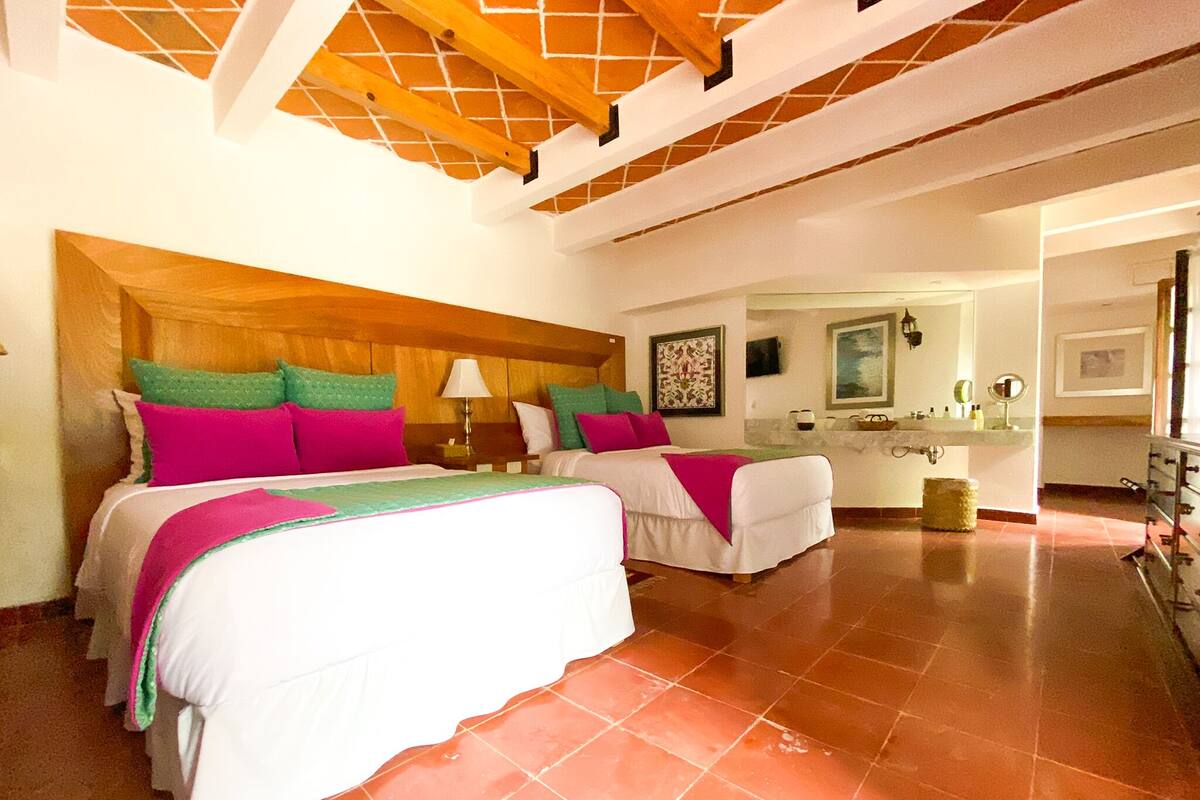 A hotel room with two beds, a wooden ceiling, a bathroom with a sink and mirror, and a tiled floor.