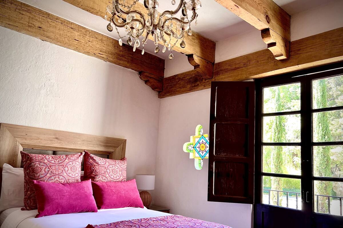 A bedroom with a wooden headboard, a bed with pink and white bedding, a chandelier, and a window with a view of greenery.