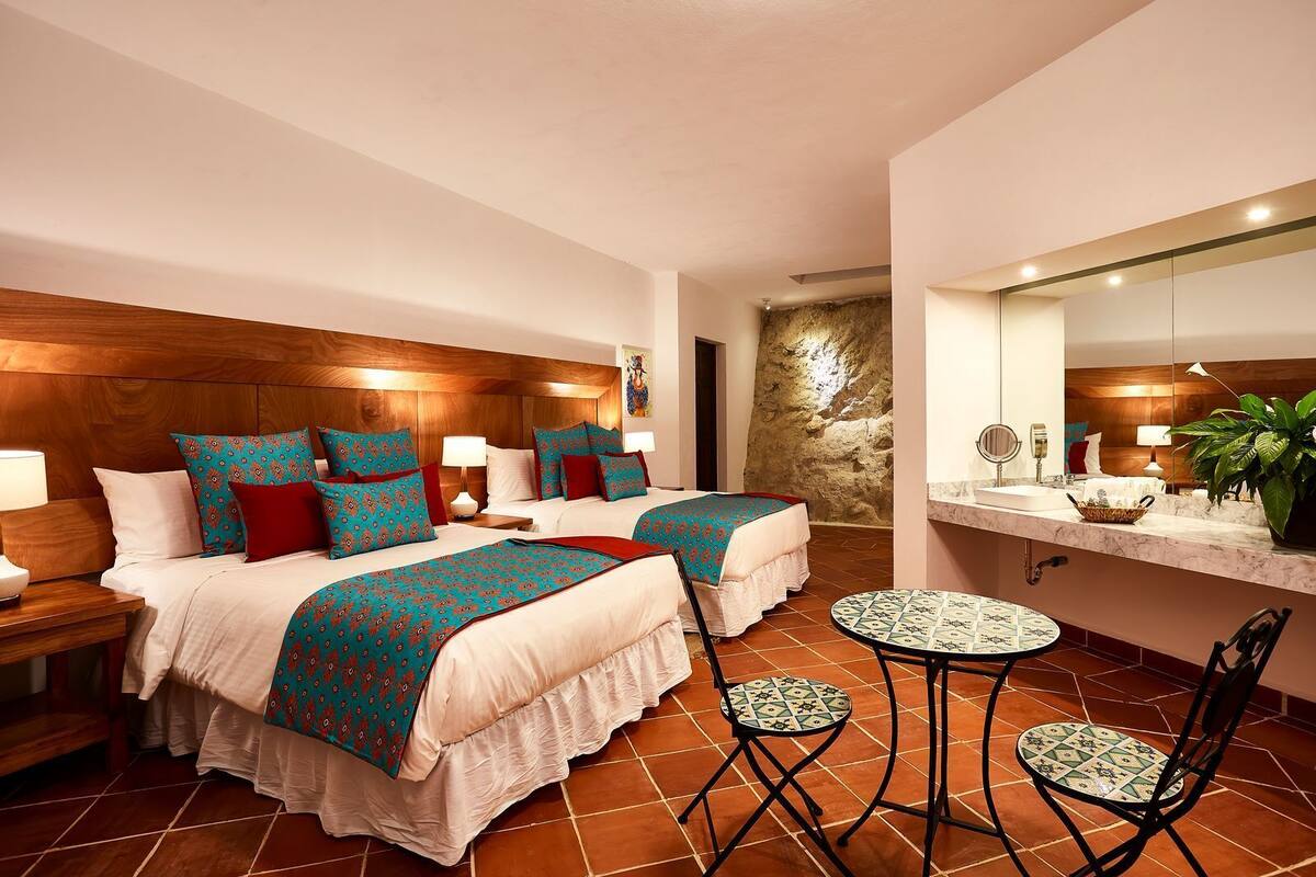A hotel room with two beds, a small round table, a chair, a bathroom with a mirror, and a stone wall.
