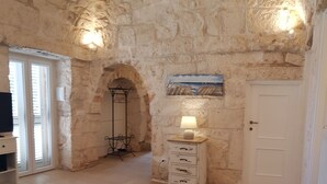 A stone-walled room with a wooden bench, a small table, and a lamp.