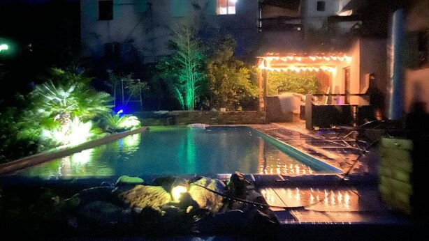 Una piscina con borde de piedra, rodeada de plantas y una pérgola con luces colgantes.