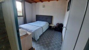 Una habitación pequeña y funcional con una cama, una mesita y una silla. Se ve una ventana en un lado y la puerta en el otro.