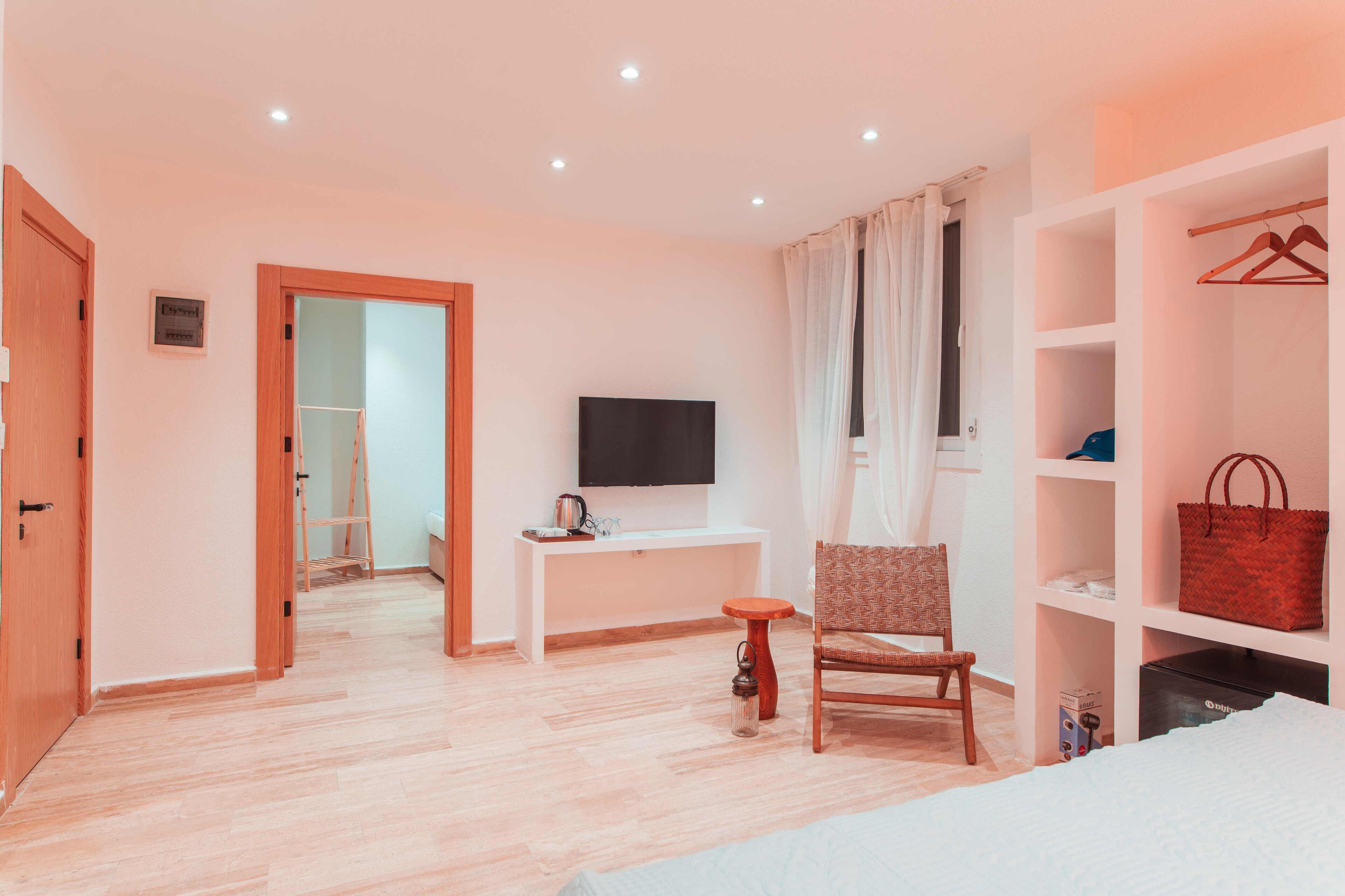 Une chambre d’hôtel avec un plancher en bois, un téléviseur posé sur un meuble blanc, une chaise, une petite table et une armoire contenant un sac rouge.