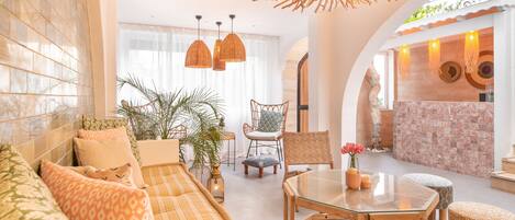 A spacious living area with a sofa, chairs, a glass coffee table, and a dining table. There is a large window with curtains, a plant, and a decorative wall with woven elements.
