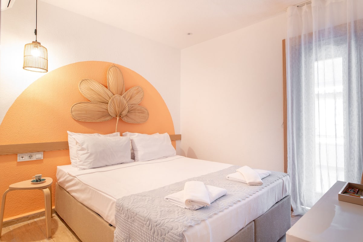 A hotel room with a bed, a small table, a chair, a desk, and a large window with curtains.