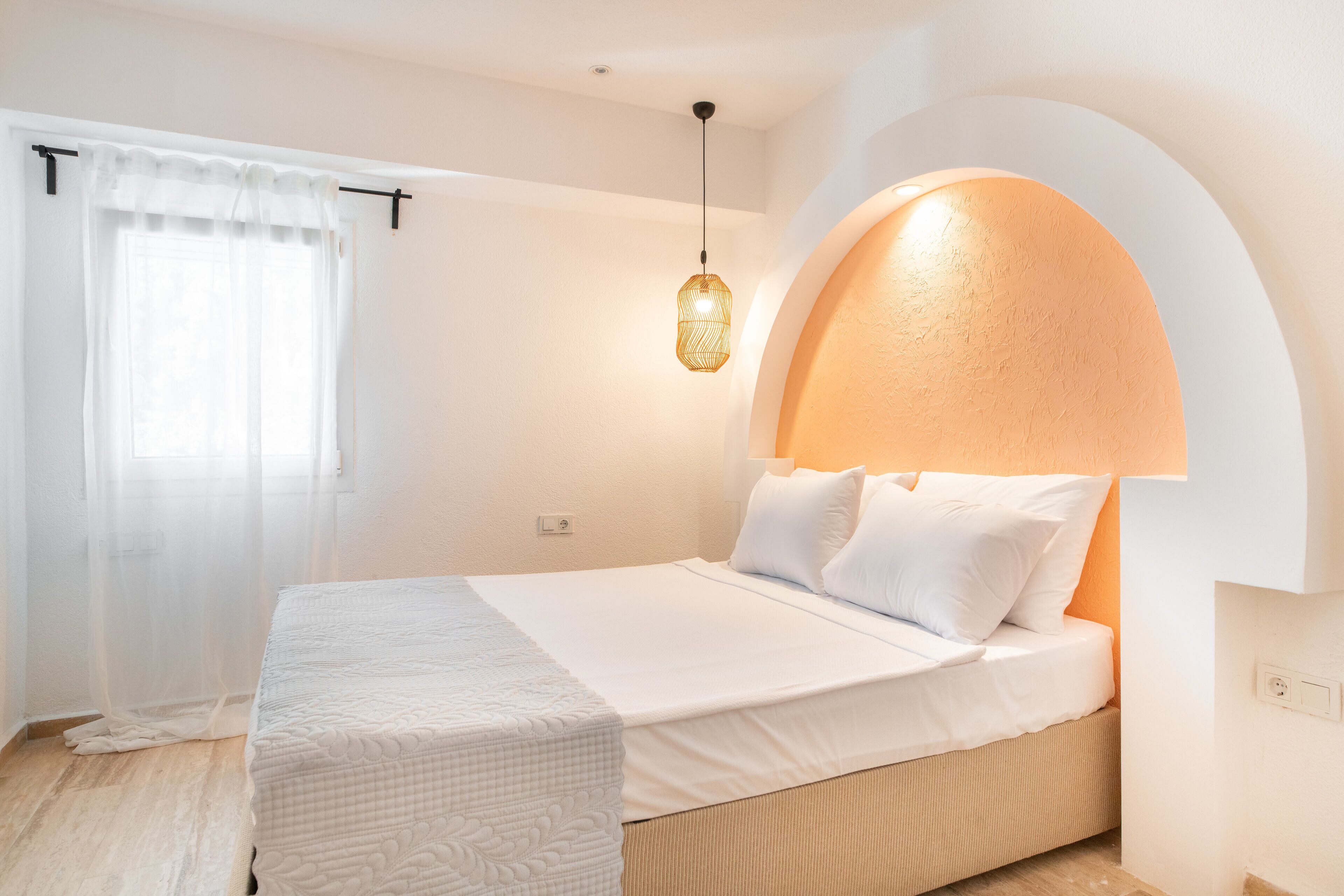 A neatly arranged bedroom with a bed, a bedside table, a hanging light fixture, and a window with sheer curtains.