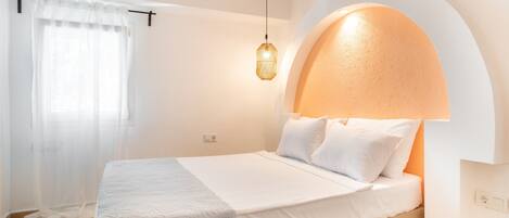 A neatly arranged bedroom with a bed, a bedside table, a hanging light fixture, and a window with sheer curtains.