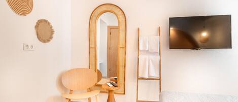 A hotel room with a wooden chair, a round mirror, a television, a towel rack, and a bed with a white comforter.