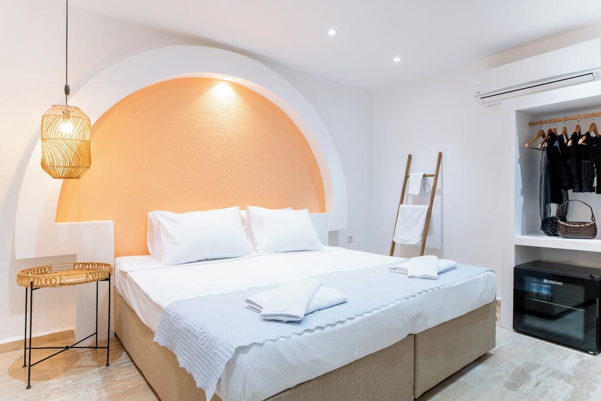 A hotel room with a bed, a bedside table, a hanging light fixture, a wardrobe, and an air conditioning unit.