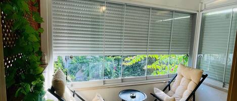 A sunlit room with a ceiling fan, large windows with blinds, two lounge chairs, a small table, and a potted plant.