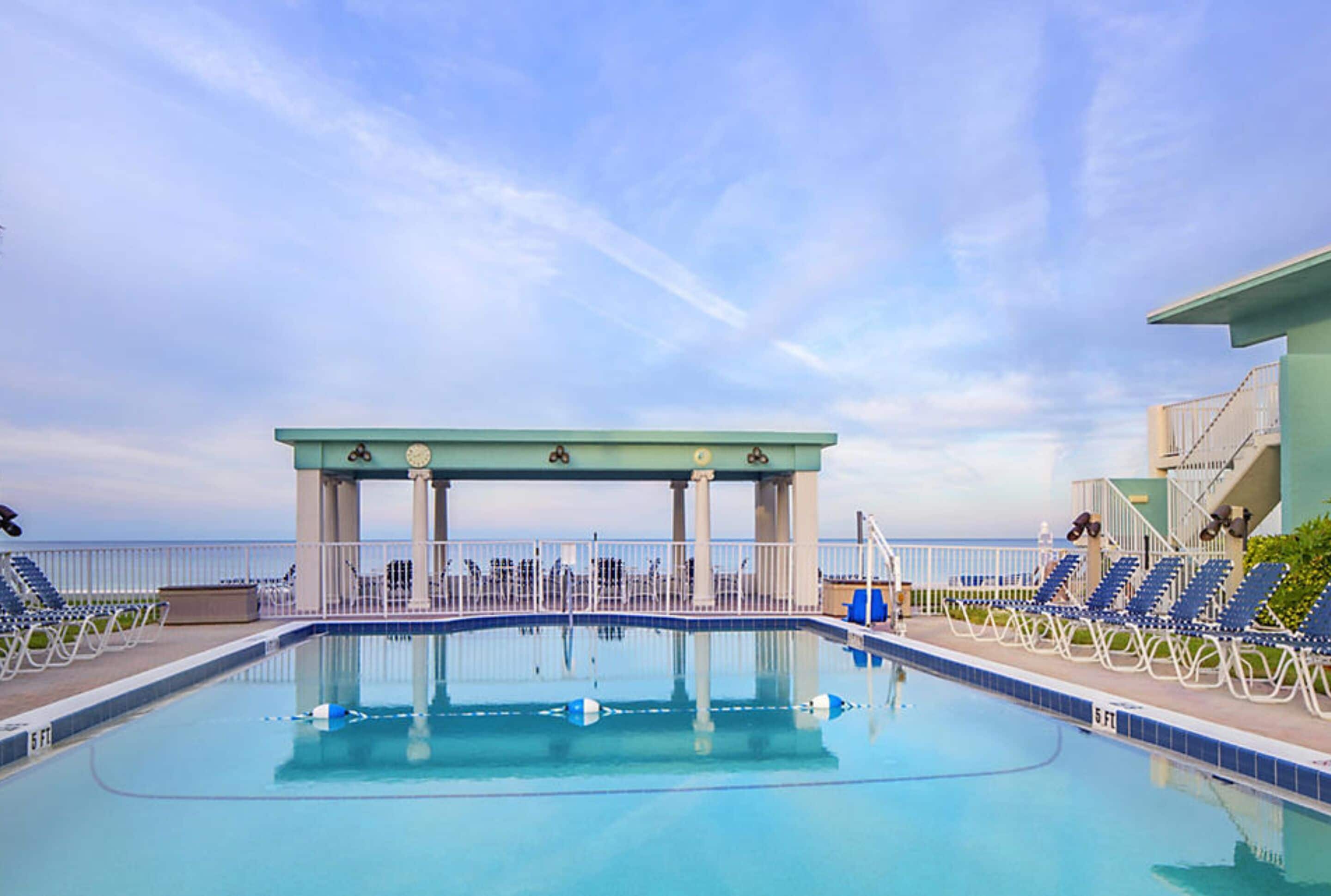 Piscina com área coberta e espreguiçadeiras, cercada por um muro branco, com vista para o oceano.