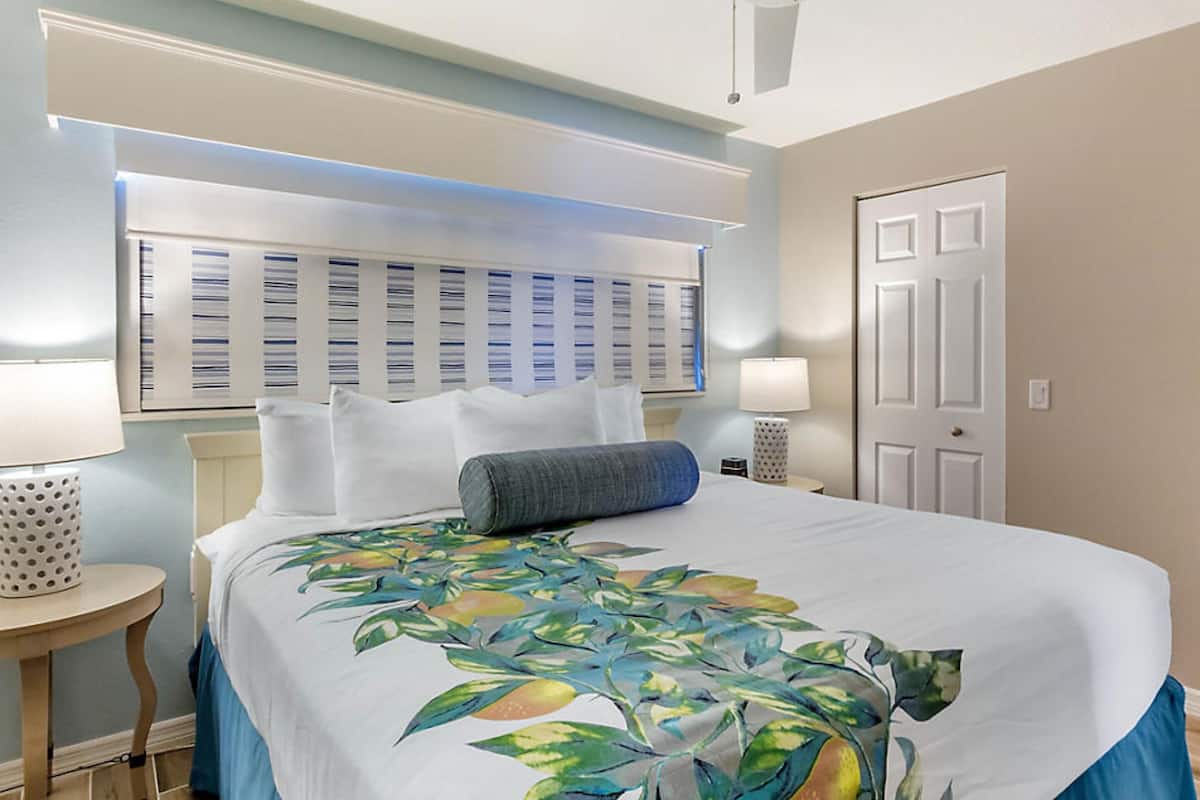 A hotel room with a large bed, a headboard with a lighted panel, a bedside table with a lamp, and a door.