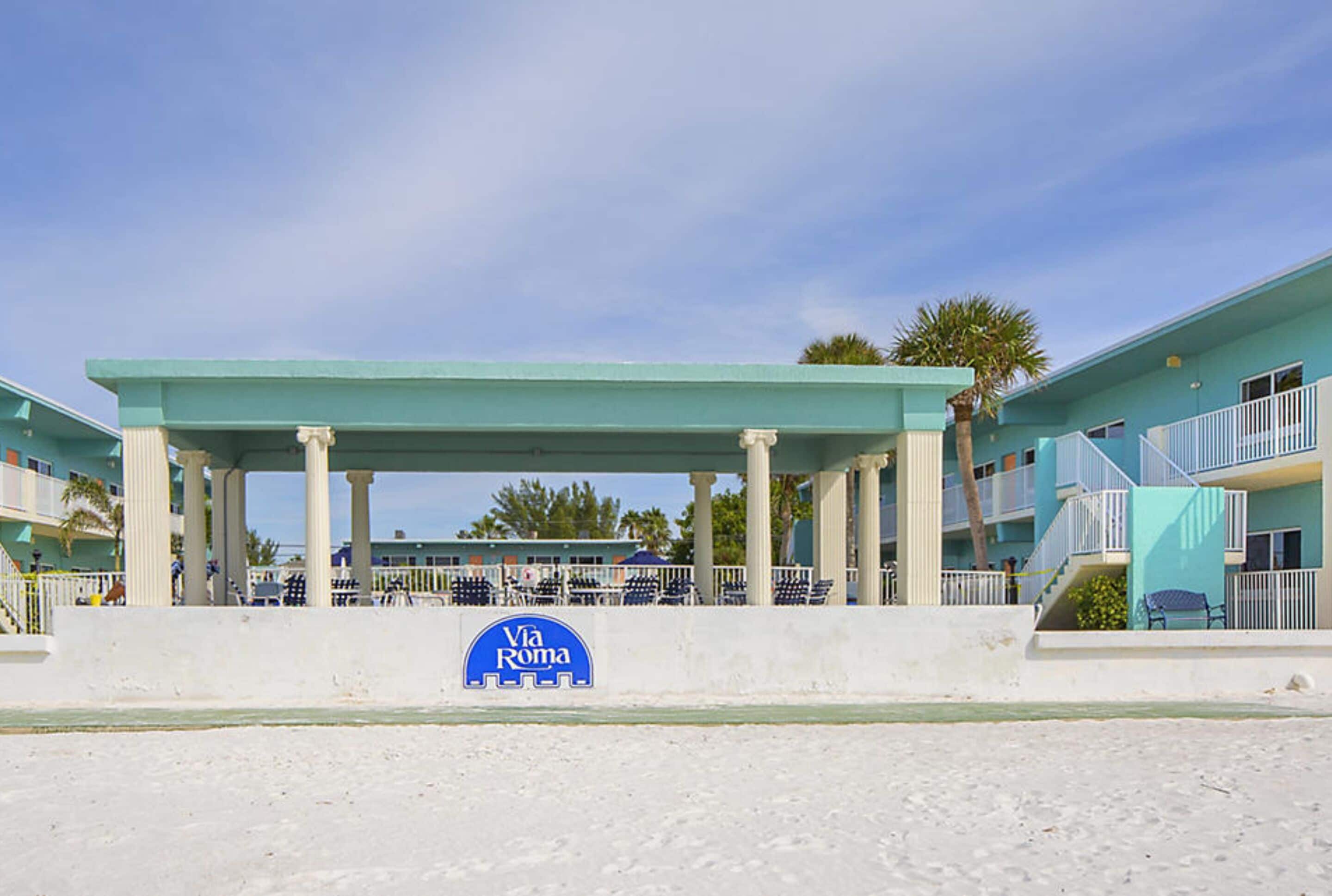 A beachside hotel with a covered outdoor seating area, a sign reading "Via Roma," and a palm tree in the background.