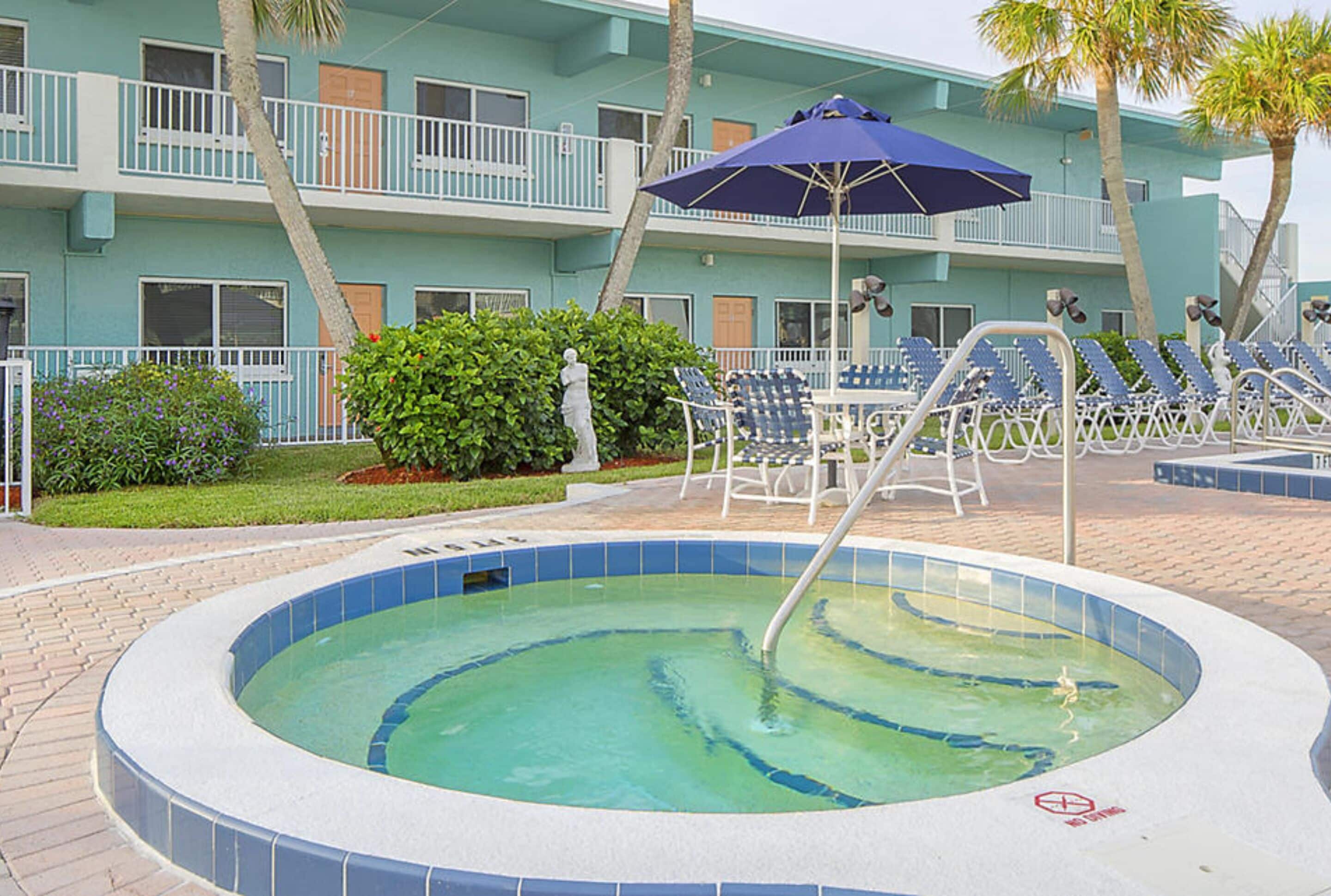 A swimming pool with lounge chairs and an umbrella, surrounded by a building with balconies and palm trees.