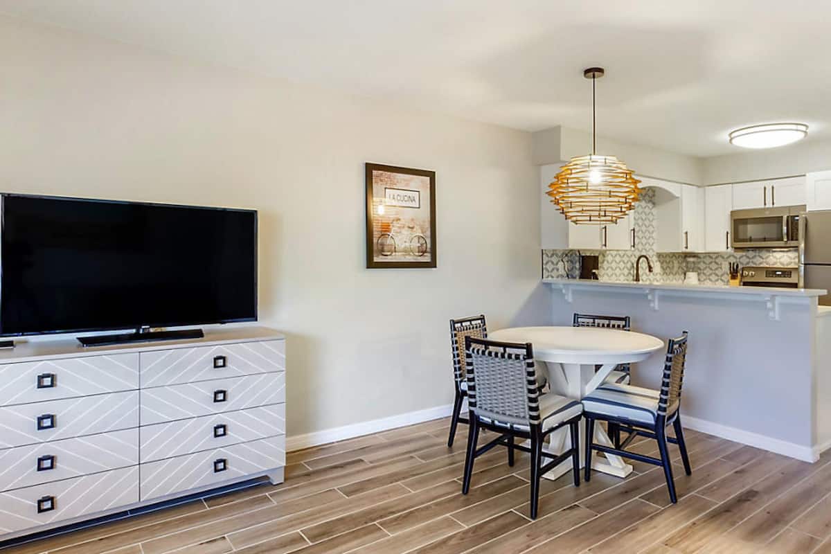 A modern kitchen with a dining area, featuring a flat-screen TV, a dining table with chairs, and a pendant light.