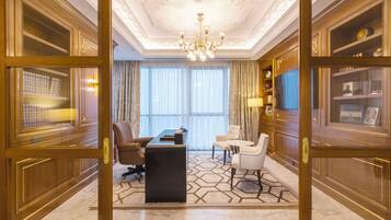 A luxurious hotel room with a large desk, two chairs, a chandelier, and a view of the city through large windows.