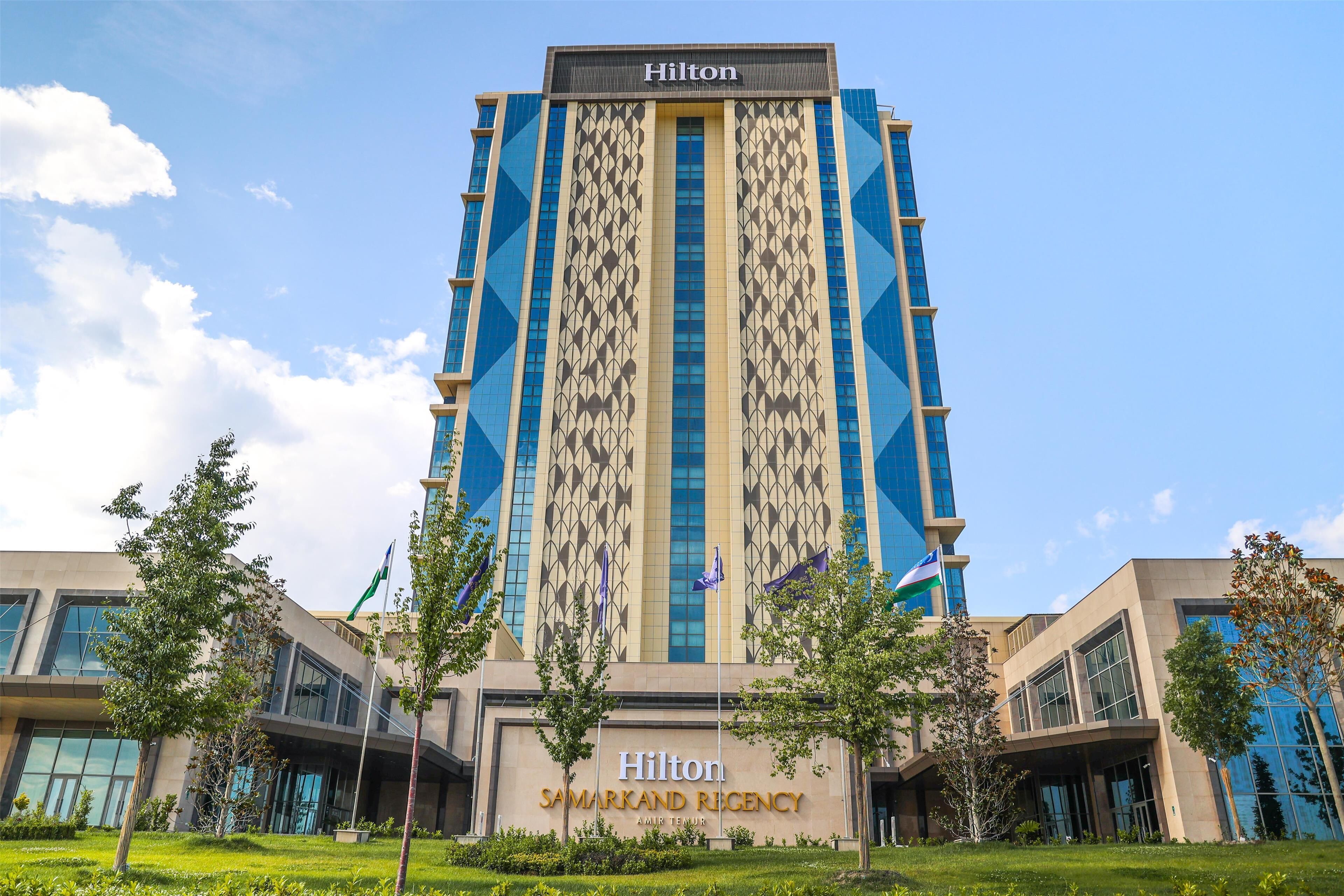 A tall, modern hotel building with a prominent Hilton logo and the name 'Samarkand Regency' displayed on the facade.