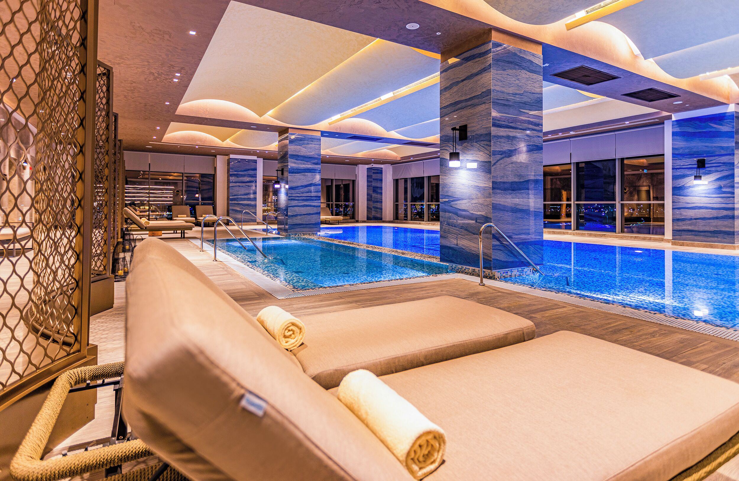 A modern indoor pool area with lounge chairs and a view of the city at night.