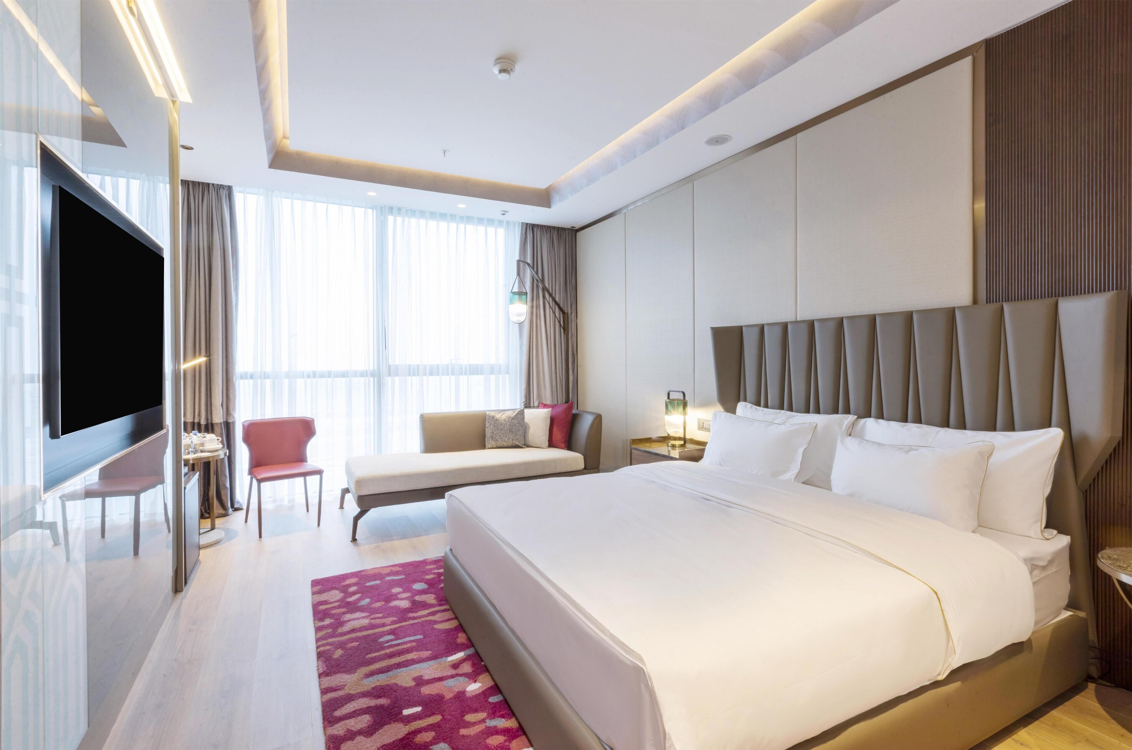 Una habitación de hotel moderna con una cama grande, un televisor de pantalla plana, una zona de estar con una silla y un sofá, y un ventanal con cortinas.