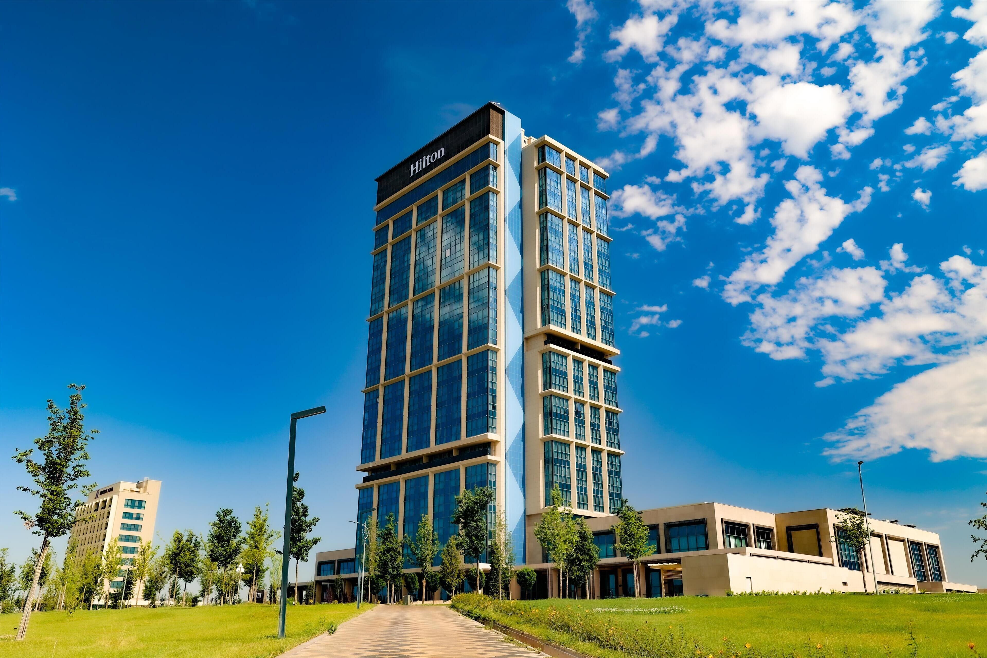 A modern hotel with a prominent 'Hilton' sign, surrounded by greenery and other buildings.