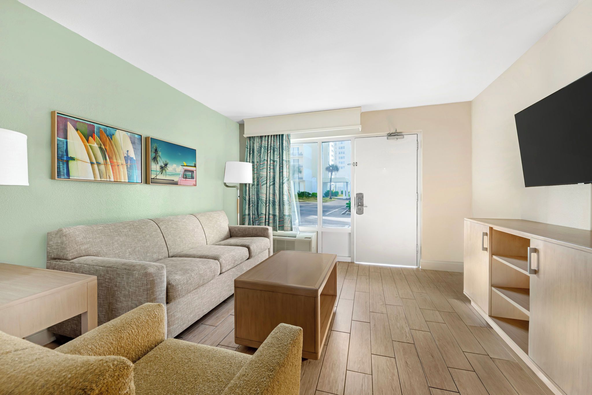 A hotel room with a sofa, armchairs, a coffee table, a TV, and a door leading to a balcony.