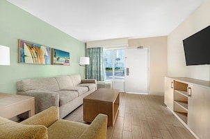 Quarto de hotel com sofá, poltronas, mesa de centro, televisão e porta para uma varanda.