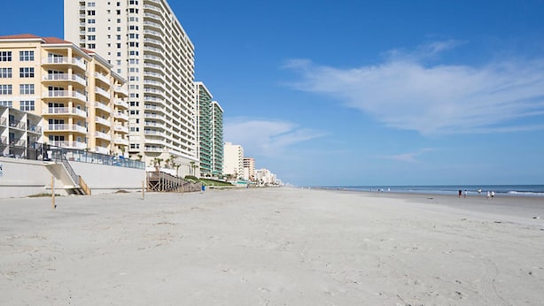 Un hotel frente a la playa con varios pisos, una playa al fondo y cielo despejado.