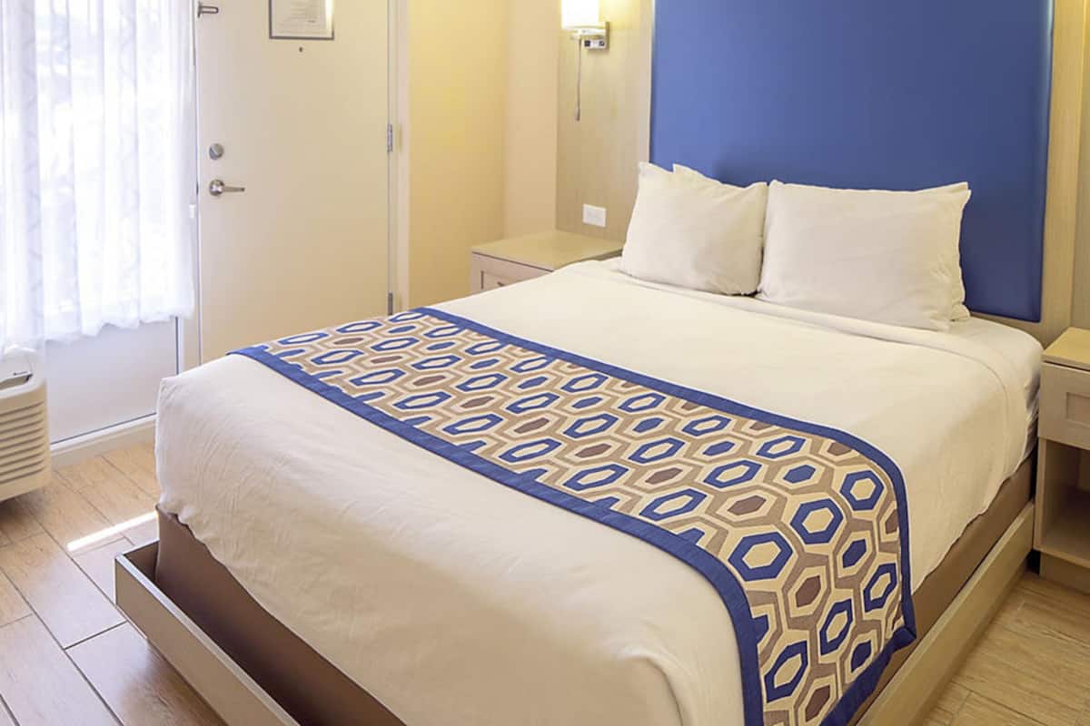 A hotel room with a bed, a nightstand, a bedside table, a window with curtains, and a wall-mounted light.