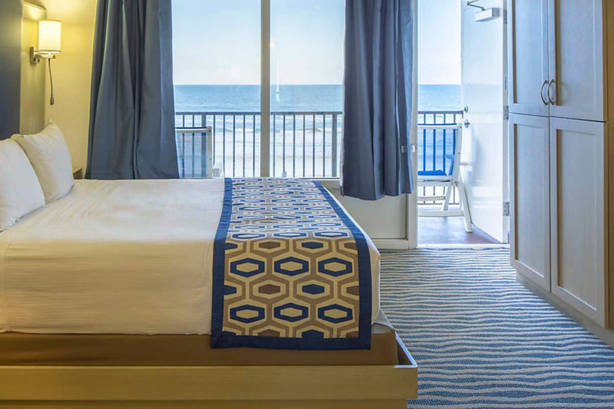 A hotel room with a large bed, a view of the ocean, a balcony with a chair, and a wardrobe.