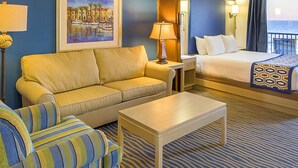 Una habitación de hotel con sofá, un sillón a rayas, cama, mesita de noche, lámpara y un cuadro con una escena costera.