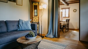 A living room with a sofa, a round wooden coffee table with a bowl of fruit, a clock, and a dining area in the background.