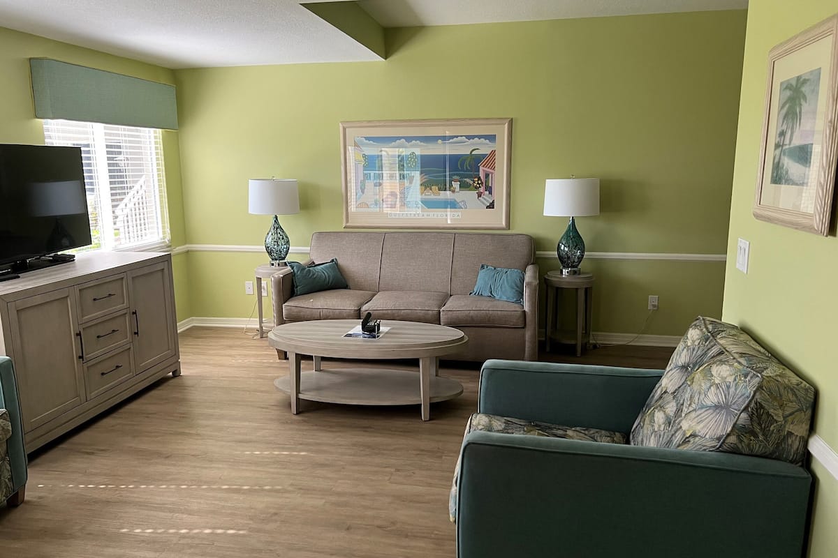 A living room with a sofa, a coffee table, and a TV on a cabinet. There are two lamps on side tables and a painting on the wall.
