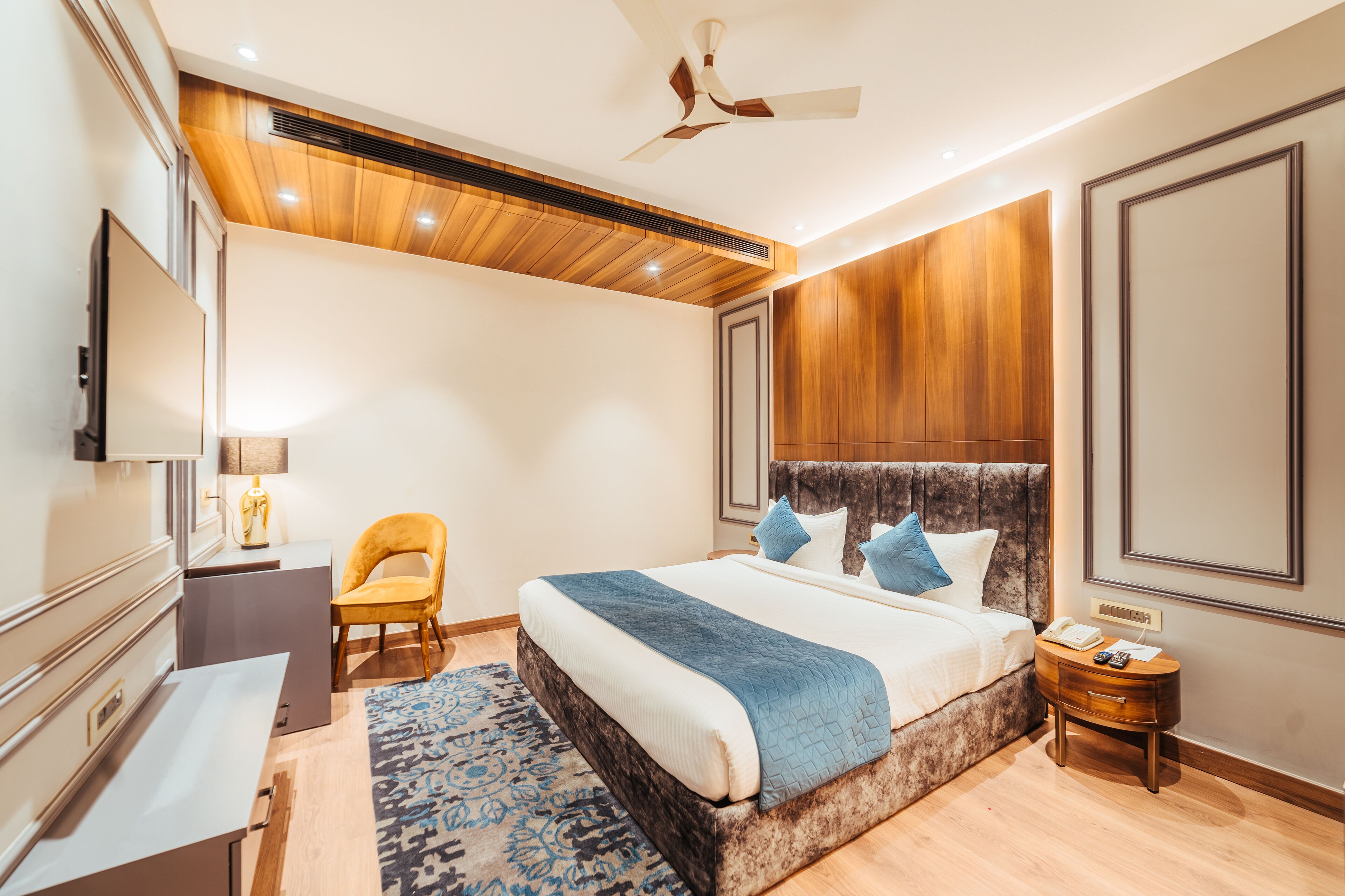 Un dormitorio moderno con una cama grande, un ventilador de techo, techo de madera, un televisor, una mesita de noche y una silla.