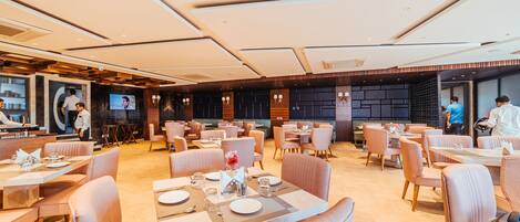 A spacious restaurant with multiple dining tables and chairs, a modern ceiling design, and a television screen on the wall.