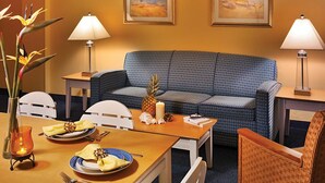 A hotel room with a blue patterned sofa, a wooden dining table set with plates, a pineapple, and a candle. There are two lamps on either side of the sofa and a vase with flowers on the left side of the table. - Fantasy Island II (Daytona Beach Shores)