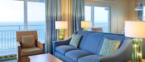 A hotel room with a large window offering a view of the sea, a blue sofa with patterned cushions, a wicker armchair, and a wooden coffee table.