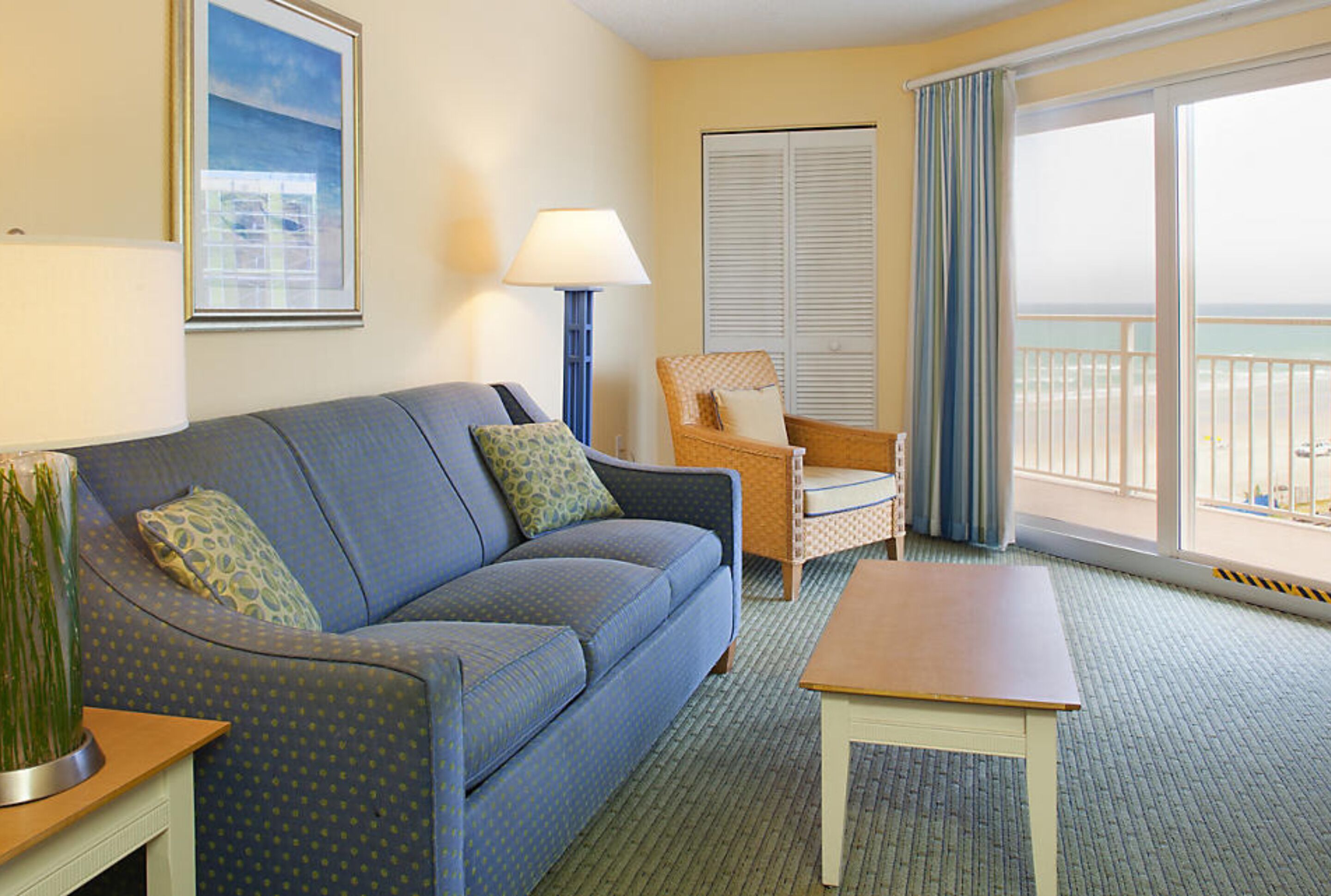 A hotel room with a blue sofa, a wooden coffee table, a wicker chair, a lamp, and a view of the beach through large windows.